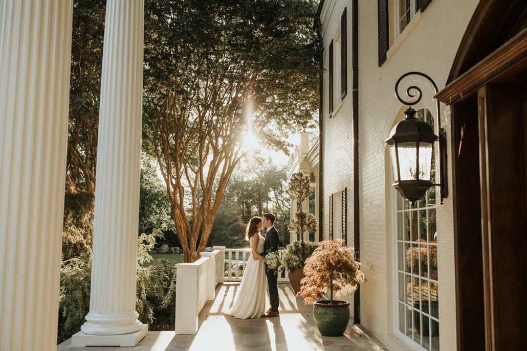 Bride and Groom standing on mansion's front veranda as sun beams through the trees behind them
