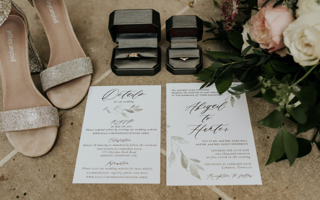 Wedding invitation suite flat lay with wedding rings, silver jeweled bridal shoes, and bouquet