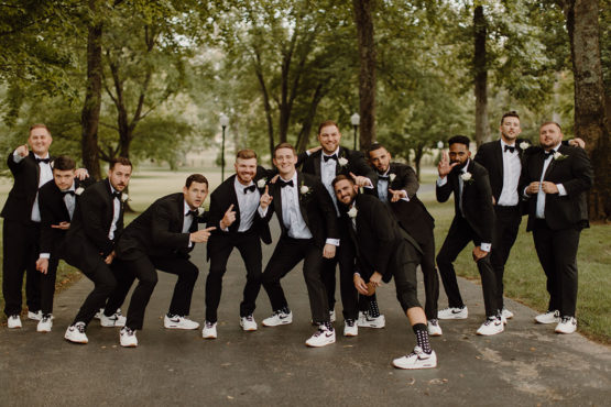 Groomsmen pose in mansion's driveway in matching classic black tuxedos and sneakers