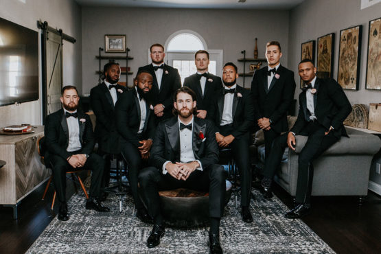 Groom poses with groomsmen, all seated in classic black tuxedos