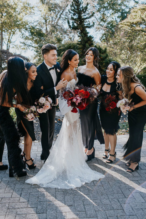 Bride in long lace gown poses and laughs with bridal crew dressed in different black outfits