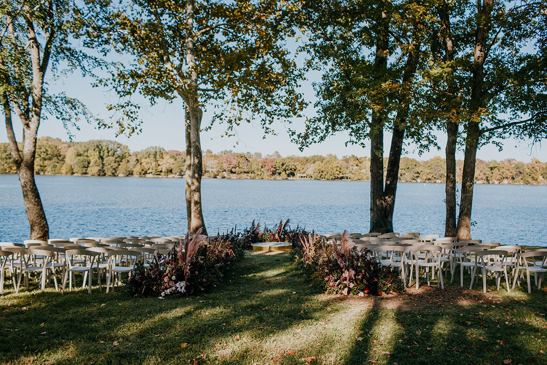 lakeside wedding ceremony with white seating and large abundant boho floral arrangements lining the aisle