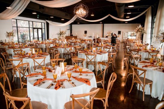 Wedding reception inside Lakeview Event Center with rust goblets and napkins on white linens with wooden crossback chairs