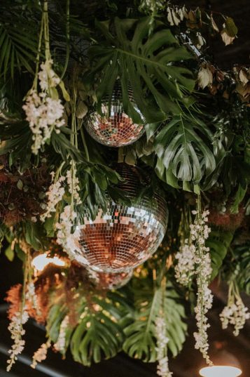 Ceiling installment of disco balls, hanging white flowers, monstera leaves, and tropical ferns