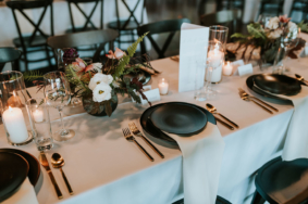Moody, modern wedding tablescape with black dishes, gold flatware, and small arrangements with fern leaves
