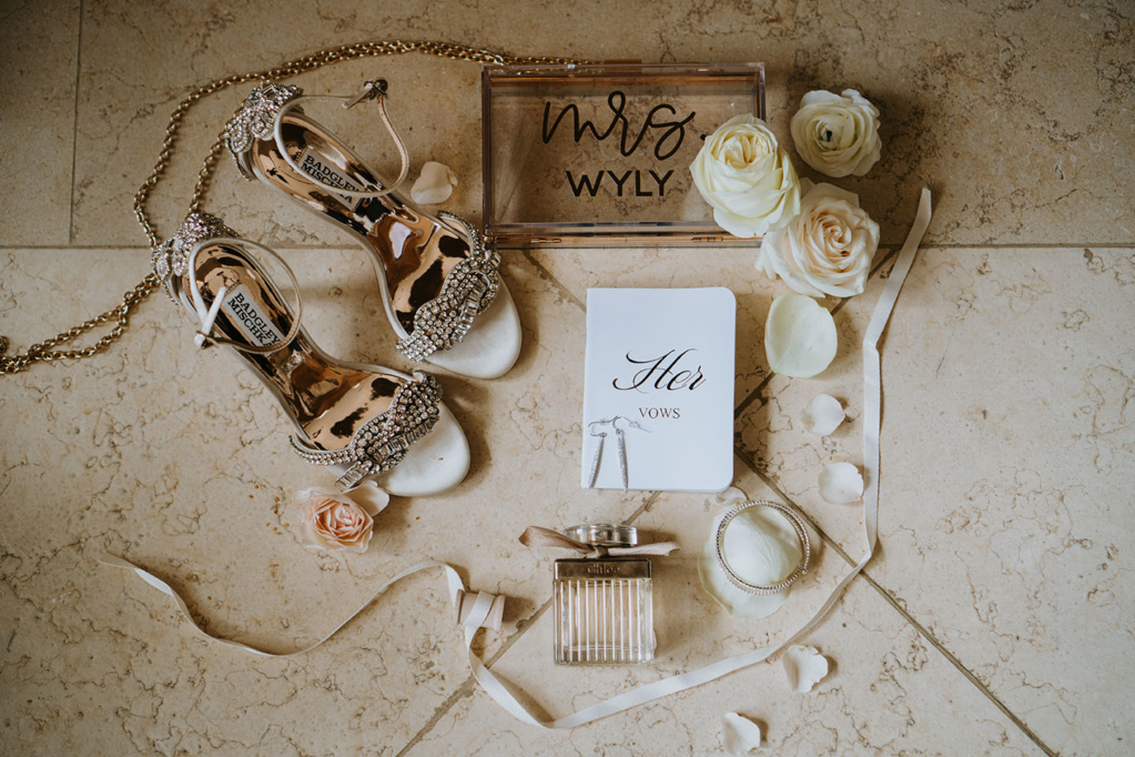Flat Lay of Wedding Day Details with jeweled bridal heels, perfume, white roses, acrylic purse