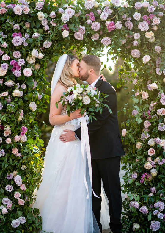 bride and groom embrace under Large Floral Ceremony Arch Backdrop with Purple Flowers