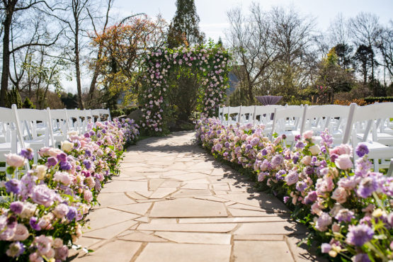 Large Floral Ceremony Arch Backdrop with Purple Flowers Ceremony Aisle Lined with Greenery and Purple Roses