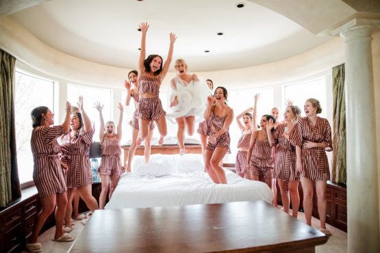 Bride cheering and jumping on Reba Suite bed with bridesmaids