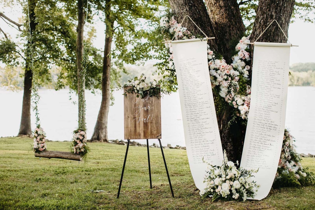 Summer Wedding Ceremony Seating Chart with elegant wooden sign and large scroll hanging from tree decorated with white and pink roses