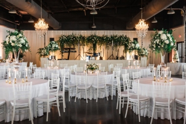 All white wedding reception with greenery arrangements and stage inside Lakeview Event Center