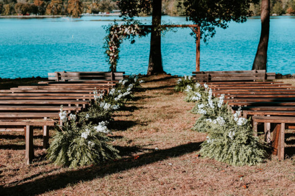 Wedding ceremony setup on the Lakeside Lawn with wooden benches, aisle lined with ferns and white flowers, and a wooden arch backdrop
