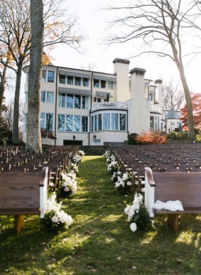 Ceremony setup on Lakeside Lawn with wooden pews and white aisle arrangements with mansion in background