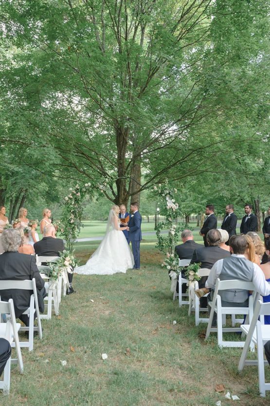 Ceremony in the willow oak canopy with white chairs and a greenery arch