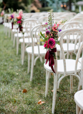 Aisle markers with bold, bright flowers and maroon ribbons on white chairs